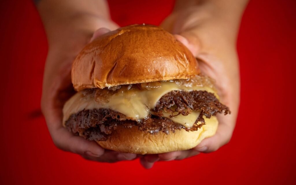 Manos sosteniendo una smash burger con queso fundido y carne caramelizada sobre un fondo rojo intenso